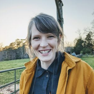 Headshot of Jessica Meyerson, a white woman with brown hair and sideswept bangs, wearing a navy blue shirt and mustard yellow overshirt. She is standing outside in front of a grassy field, with trees in the background.